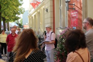 Interakcję między mówcą a słuchaczami. Przed budynkiem "Rada Miejska w Oławie" zebrała się grupa ludzi, by wysłuchać mężczyzny z mikrofonem. Kontekst wskazuje na oficjalne wydarzenie lub spotkanie dotyczące spraw miejskich.