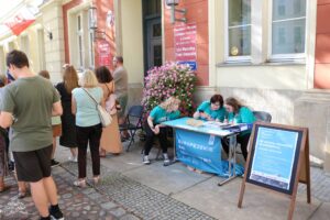 Grupa osób stoi przed budynkiem z polską flagą. Przy stole z niebieskim banerem "EUROPEJSKIE DNI DZIEDZICTWA" stoją trzy kobiety w tealowych koszulkach. Drewniany znak A-frame przed stołem zapowiada wydarzenie poświęcone architekturze Oławy, które odbędzie się 20 września 2025 roku.