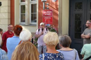 Interakcję między mówcą a słuchaczami. Przed budynkiem "Rada Miejska w Oławie" zebrała się grupa ludzi, by wysłuchać mężczyzny z mikrofonem. Kontekst wskazuje na oficjalne wydarzenie lub spotkanie dotyczące spraw miejskich.