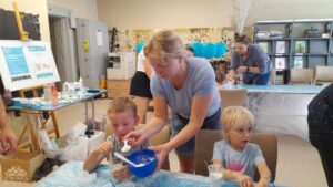 Dzieci siedzą za stołami z niebieską ceratą, zajęte warsztatami plastycznymi.