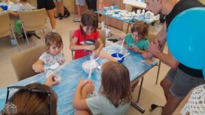 Dzieci siedzą za stołami z niebieską ceratą, zajęte warsztatami plastycznymi.