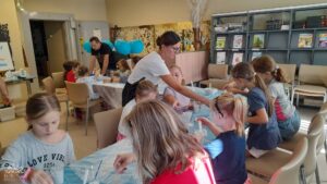 Dzieci siedzą za stołami z niebieską ceratą, zajęte warsztatami plastycznymi.