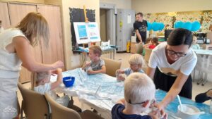 Dzieci siedzą za stołami z niebieską ceratą, zajęte warsztatami plastycznymi.