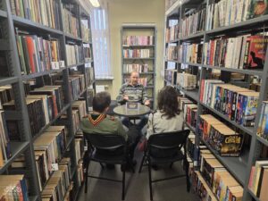 Opis wygenerowany automatycznie: Trzy osoby siedzą przy małym okrągłym stole w bibliotece, otoczone wysokimi regałami z książkami. Starszy mężczyzna w swetrze rozmawia z dwoma młodymi ludźmi w mundurach harcerskich.