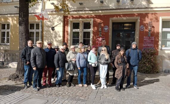 Opis wygenerowany automatycznie: Grupa około piętnastu osób, ubranych w jesienne kurtki i czapki, stoi na brukowanym placu przed zabytkowym, dwupiętrowym budynkiem o czerwono-pomarańczowej fasadzie i białych oknach. Na budynku wisi polska flaga. Po lewej stronie zdjęcia widoczne jest duże drzewo.