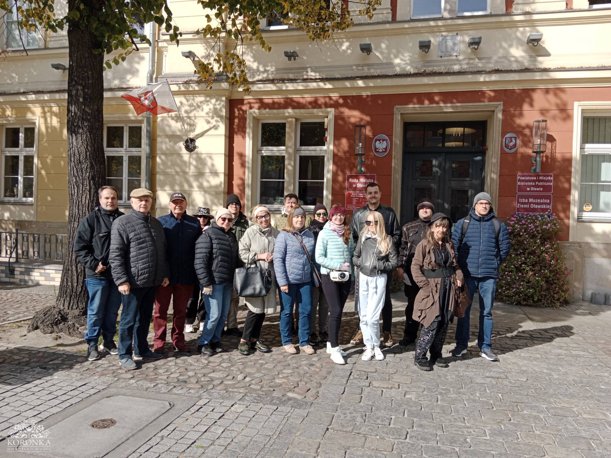Opis wygenerowany automatycznie: Grupa około piętnastu osób, ubranych w jesienne kurtki i czapki, stoi na brukowanym placu przed zabytkowym, dwupiętrowym budynkiem o czerwono-pomarańczowej fasadzie i białych oknach. Na budynku wisi polska flaga. Po lewej stronie zdjęcia widoczne jest duże drzewo.