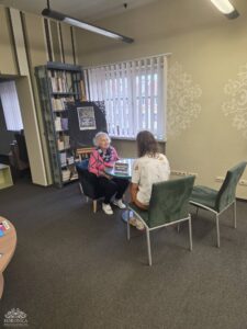 Opis wygenerowany automatycznie: Dwie kobiety siedzą naprzeciwko siebie przy szklanym stoliku w jasnym pomieszczeniu, prawdopodobnie czytelni. Starsza kobieta w różowym swetrze i młodsza kobieta w białej koszuli rozmawiają. W tle widać okno z żaluzjami i regał z książkami.