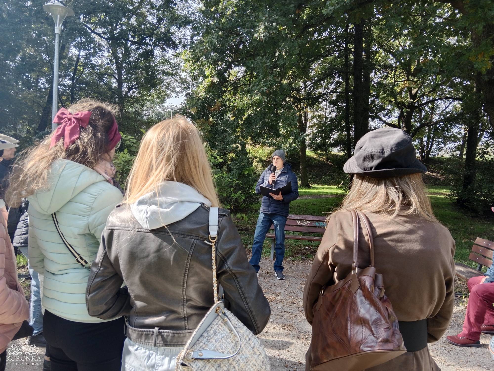 Opis wygenerowany automatycznie: Trzy osoby, widziane od tyłu, słuchają mężczyzny stojącego przed nimi na ścieżce w parku. Mężczyzna trzyma mikrofon i teczkę, a za nim widać gęste drzewa i ławki parkowe. Słuchacze są ubrani w jesienne kurtki.