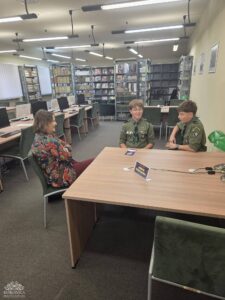 Opis wygenerowany automatycznie: Trzy osoby siedzą przy dużym drewnianym stole w przestronnej bibliotece. Kobieta w kolorowej kwiecistej marynarce rozmawia z dwoma młodymi chłopcami w mundurach harcerskich. W tle widać rzędy biurek z komputerami i regały z książkami.