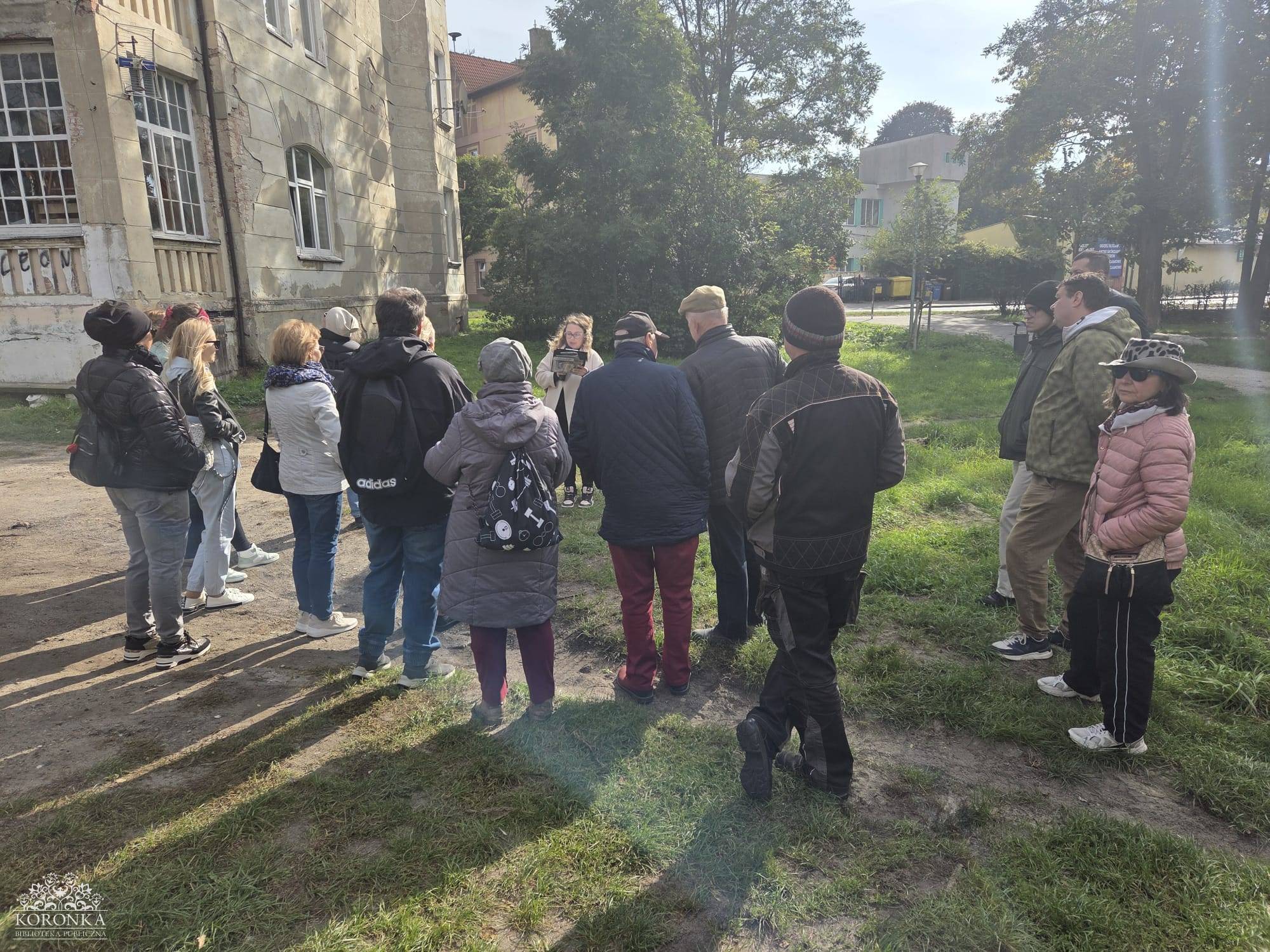 Opis wygenerowany automatycznie: Grupa około dwunastu osób stoi na trawiastym terenie, otaczając jedną osobę, która wydaje się być przewodnikiem lub prelegentem. W tle widać fragment starego budynku i drzewa. Scena rozgrywa się na zewnątrz, w słoneczny dzień.