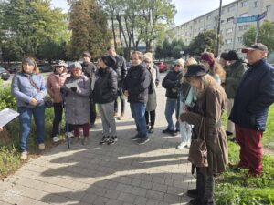 Opis wygenerowany automatycznie: Grupa dorosłych ludzi stoi na chodniku w miejskim otoczeniu, otoczona drzewami i budynkami mieszkalnymi. Większość z nich jest ubrana w kurtki i czapki, a niektórzy trzymają w rękach dokumenty, wyglądając, jakby słuchali przewodnika lub uczestniczyli w zorganizowanym spotkaniu.