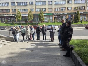 Opis wygenerowany automatycznie: Grupa dorosłych ludzi stoi na chodniku przed rzędem budynków z lokalami usługowymi na parterze. W tle widać ulicę z zaparkowanymi samochodami. Ludzie są ubrani w codzienne ubrania i wydają się być zgromadzeni w celu słuchania lub obserwowania czegoś.