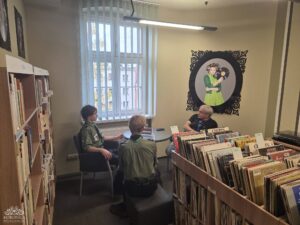 Opis wygenerowany automatycznie: Trzy osoby – dwóch młodych ludzi w mundurach harcerskich i starszy mężczyzna – siedzą wokół małego okrągłego stołu w przytulnym kąciku biblioteki. W tle widać regały z książkami i płytami, a na ścianie wisi charakterystyczny obraz kobiety z wałkami na włosach trzymającej płytę winylową. Obok stołu znajduje się okno z żaluzjami.