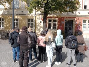 Opis wygenerowany automatycznie: Grupa dorosłych ludzi stoi na brukowanym placu, tyłem do widza, patrząc w kierunku drzewa i zabytkowych budynków w tle. Są ubrani w codzienne ubrania, a scena sugeruje zorganizowane spotkanie lub wycieczkę.