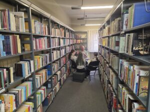 Opis wygenerowany automatycznie: Cztery osoby, w tym jeden mężczyzna i trzy młode kobiety, siedzą wokół małego stołu w wąskim korytarzu biblioteki, otoczone wysokimi regałami pełnymi książek. Wydają się prowadzić rozmowę lub spotkanie.