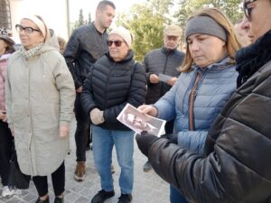 Opis wygenerowany automatycznie: Grupa ludzi stoi na zewnątrz, a kobieta w prawym dolnym rogu trzyma małe czarno-białe zdjęcie mężczyzny, które pokazuje innym.