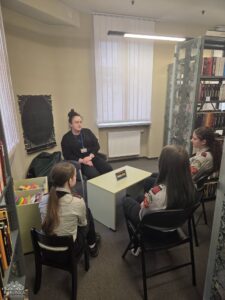 Opis wygenerowany automatycznie: Cztery osoby, w tym jedna dorosła kobieta i trzy młode kobiety, siedzą wokół jasnego stołu w rogu biblioteki. Dorosła kobieta patrzy w stronę kamery, a pozostałe trzy osoby są zwrócone w jej stronę, prowadząc rozmowę.