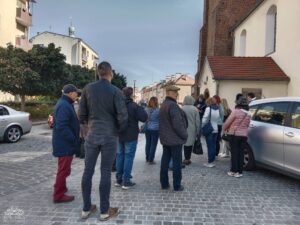 Opis wygenerowany automatycznie: Grupa ludzi stoi na brukowanej ulicy, większość odwrócona plecami do kamery, patrząc w kierunku budynku z ceglaną fasadą po prawej stronie.