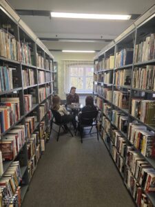 Opis wygenerowany automatycznie: Trzy osoby, w tym jedna dorosła kobieta i dwie młode kobiety, siedzą wokół małego, okrągłego stołu w środku długiego korytarza bibliotecznego, otoczonego regałami z książkami. W tle widać okno z żaluzjami.