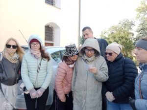 Opis wygenerowany automatycznie: Grupa ludzi stoi na zewnątrz, a kobieta w jasnym płaszczu z kapturem w centrum patrzy na kartkę papieru, którą trzyma w rękach.