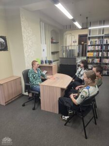 Opis wygenerowany automatycznie: Kobieta w zielonej koszuli w kropki siedzi przy biurku i rozmawia z trzema młodymi osobami w mundurach, prawdopodobnie harcerskich, w pomieszczeniu przypominającym bibliotekę lub biuro.