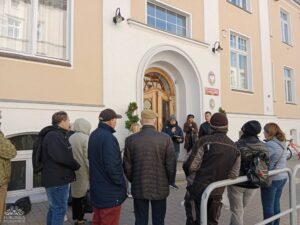 Opis wygenerowany automatycznie: Grupa około dziesięciu osób stoi na chodniku przed jasnym budynkiem z dużym łukowatym wejściem. Jedna osoba w środku grupy, ubrana w ciemną kurtkę i czapkę, wydaje się przemawiać do pozostałych, trzymając w ręku małe urządzenie. Na ścianie budynku, po prawej stronie wejścia, wisi czerwono-biały herb. Ludzie są ubrani w ciepłe, codzienne ubrania.