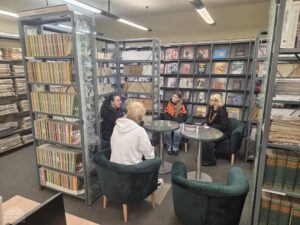 Opis wygenerowany automatycznie: Cztery osoby siedzą w wygodnych fotelach wokół dwóch małych stolików w bibliotece, otoczone regałami z książkami i czasopismami, prowadząc swobodną rozmowę.