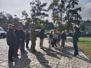 Opis wygenerowany automatycznie: Grupa około piętnastu osób stoi na brukowanej ścieżce na zewnątrz, słuchając osoby stojącej po prawej stronie kadru, która wydaje się do nich przemawiać. W tle widać duże drzewa z zielonymi liśćmi, zaparkowane samochody i inne budynki. Słońce rzuca długie cienie na bruk.