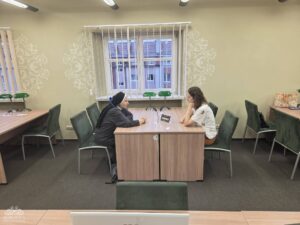 Opis wygenerowany automatycznie: Dwie kobiety siedzą naprzeciwko siebie przy drewnianym stole; jedna ubrana jest w ciemny habit, a druga w jasną koszulkę, rozmawiając w pokoju z dużym oknem i ozdobnymi ścianami.