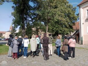 Opis wygenerowany automatycznie: Duża grupa ludzi, widziana głównie od tyłu, stoi na brukowanym placu, patrząc w kierunku gęstych, zielonych drzew. W tle po lewej stronie widać fragment budynku z wieżą lub iglicą, prawdopodobnie kościoła, a po prawej stronie fragment innego budynku. Niebo jest błękitne z kilkoma chmurami.