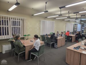 Opis wygenerowany automatycznie: Grupa osób, w tym kilka w mundurach harcerskich lub podobnych, siedzi przy drewnianych stołach w jasnym pomieszczeniu przypominającym bibliotekę lub czytelnię.