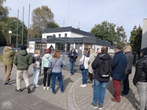 Opis wygenerowany automatycznie: Grupa dorosłych ludzi stoi na brukowanym placu przed nowoczesnym budynkiem z ciemnym dachem, słuchając mężczyzny trzymającego książkę lub notatki, który do nich przemawia. W tle widać drzewa i ogrodzenie.