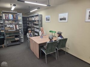 Opis wygenerowany automatycznie: Kobieta w jasnej bluzce siedzi przy stole z trzema młodymi osobami w mundurach, prawdopodobnie harcerskich, w pomieszczeniu z regałami pełnymi książek.