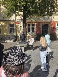 Opis wygenerowany automatycznie: Grupa ludzi, w tym dzieci i dorośli, stoi na brukowanym placu pod dużym drzewem, obserwując dwoje młodych ludzi (mężczyznę i kobietę) stojących przy kamiennym stole. Kobieta trzyma mikrofon lub inne urządzenie. W tle widać kolorowe budynki z oknami i polską flagę. Na pierwszym planie widoczny jest kapelusz w panterkę.