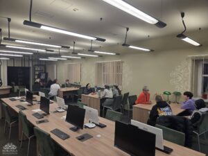 Opis wygenerowany automatycznie: Szeroki widok na dużą czytelnię lub salę komputerową, gdzie wiele osób, w tym dzieci i dorośli, niektórzy w mundurach, siedzi przy stołach. Część stołów wyposażona jest w monitory komputerowe, a inni uczestnicy rozmawiają ze sobą.