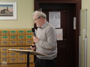 Opis wygenerowany automatycznie: Mężczyzna w okularach i jasnoszarym swetrze stoi za drewnianym pulpitem, mówiąc do mikrofonu. W tle widoczne są drewniane szafki z małymi szufladkami, oznaczonymi literami i słowami, oraz drzwi z wywieszoną informacją o godzinach otwarcia czytelni.