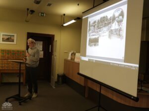 Opis wygenerowany automatycznie: Mężczyzna w okularach i jasnoszarym swetrze stoi za pulpitem, mówiąc do mikrofonu, i patrzy w stronę dużego ekranu projekcyjnego. Na ekranie wyświetlany jest stary, ilustrowany obraz, prawdopodobnie historyczna pocztówka lub mapa.