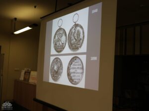 Opis wygenerowany automatycznie: Zbliżenie na ekran projekcyjny, na którym wyświetlone są dwa zestawy historycznych medali lub monet. Górny zestaw, oznaczony rokiem "1848", przedstawia awers i rewers medalu z kogutem i skrzyżowanymi narzędziami. Dolny zestaw, oznaczony rokiem "1898", również pokazuje awers i rewers medalu, w tym jeden z kogutem i drugi z tekstem.