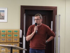 Opis wygenerowany automatycznie: Mężczyzna w okularach i bordowym swetrze stoi, trzymając mikrofon w prawej ręce. W tle widać drewniane szafki katalogowe z szufladami oraz zamknięte drzwi.