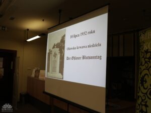 Opis wygenerowany automatycznie: Ekran projektora wyświetla slajd z tekstem "10 lipca 1932 roku", "Oławska krwawa niedziela" i "Der Ohlauer Blutsonntag". Po lewej stronie ekranu znajduje się czarno-białe zdjęcie pomnika z symbolem swastyki. Scena jest słabo oświetlona.