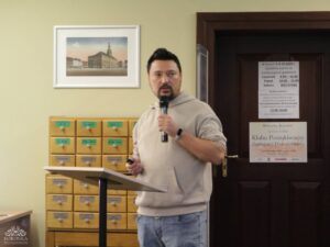 Opis wygenerowany automatycznie: Mężczyzna w jasnej bluzie z kapturem i dżinsach stoi, mówiąc do mikrofonu i trzymając w lewej ręce małe urządzenie. W tle widać drewniane szafki katalogowe, obraz na ścianie oraz drzwi z przyklejoną kartką informacyjną.