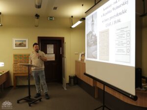 Opis wygenerowany automatycznie: Mężczyzna w jasnej bluzie i dżinsach stoi przy mównicy z mikrofonem, prezentując slajd na projektorze. Na slajdzie widoczne są teksty w języku polskim i niemieckim, dotyczące wyborów prezydenckich w Republice Weimarskiej. W tle widać regały z szufladami i półki.