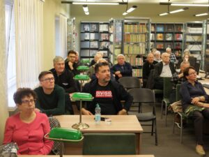 Opis wygenerowany automatycznie: Grupa ludzi, głównie dorosłych i starszych, siedzi na krzesłach w pomieszczeniu przypominającym bibliotekę lub salę wykładową. W tle widoczne są regały z książkami. Niektórzy z nich patrzą w kierunku prezentacji, inni rozglądają się. Na stołach przed nimi stoją lampki.