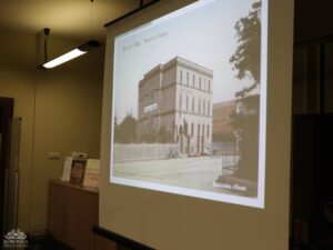 Opis wygenerowany automatycznie: Ekran projektora wyświetla stare, czarno-białe zdjęcie budynku. Na zdjęciu widnieje napis "Gruß aus Ohlau. Denkmal am Hermann." oraz "Biblioteka-Oława". Budynek ma wiele okien i wydaje się być historyczną budowlą, być może biblioteką lub księgarnią.