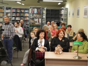 Opis wygenerowany automatycznie: Grupa ludzi, głównie dorosłych, siedzi w pomieszczeniu przypominającym bibliotekę, z regałami pełnymi książek w tle. Mężczyzna w koszuli w kratę stoi po lewej stronie, a większość osób patrzy w jednym kierunku, prawdopodobnie na prelegenta.