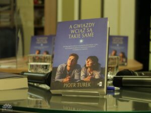 Opis wygenerowany automatycznie: Zbliżenie na książkę zatytułowaną "A GWIAZDY WCIAŻ SĄ TAKIE SAME" autorstwa Piotra Turka, stojącą na podstawce na stole. Na okładce widać mężczyznę i kobietę patrzących w górę na niebo z księżycem. W tle widać rozmyte inne egzemplarze książki, mikrofony i szklanki z wodą.