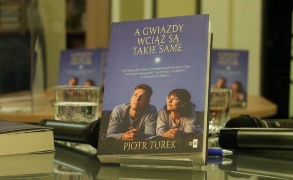 Opis wygenerowany automatycznie: Zbliżenie na książkę zatytułowaną "A GWIAZDY WCIAŻ SĄ TAKIE SAME" autorstwa Piotra Turka, stojącą na podstawce na stole. Na okładce widać mężczyznę i kobietę patrzących w górę na niebo z księżycem. W tle widać rozmyte inne egzemplarze książki, mikrofony i szklanki z wodą.