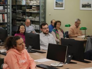 Opis wygenerowany automatycznie: Grupa dorosłych osób siedzi przy biurkach z monitorami komputerowymi, uczestnicząc w spotkaniu lub zajęciach. Kobieta w różowej bluzie z kapturem jest na pierwszym planie po lewej, a mężczyzna w okularach i z brodą siedzi pośrodku, patrząc przed siebie. W tle widoczne są regały z książkami.