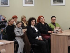 Opis wygenerowany automatycznie: Grupa kobiet i młoda dziewczynka siedzą w rzędach na widowni, patrząc w kierunku poza kadr. W tle widać ściany z oprawionymi obrazami.