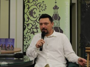 Opis wygenerowany automatycznie: Mężczyzna z brodą, ubrany w białą koszulę, siedzi i mówi do mikrofonu. Za nim znajduje się zielony baner z napisem "BIBLIOTEKA KORONKA" i adresem strony internetowej. Na stole obok niego leżą dwie książki.